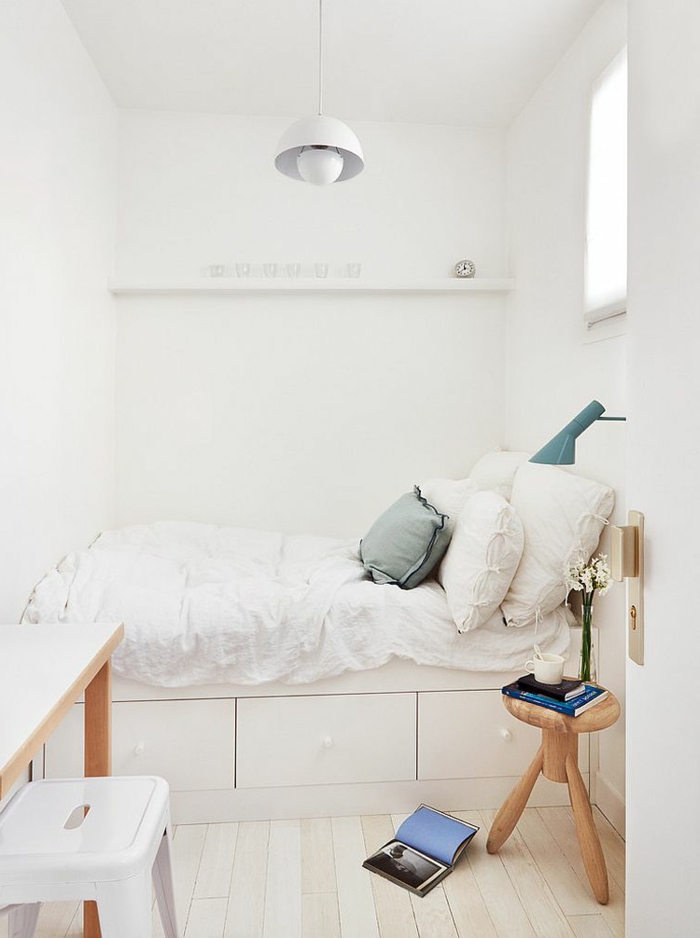 schmales Schlafzimmer in Weiß, kleines Bett mit Kasten, runder Hocker aus Hold, quadratisches Fenster über dem Betttisch, schmaler Schreibtisch mit weißem Metallstuhl